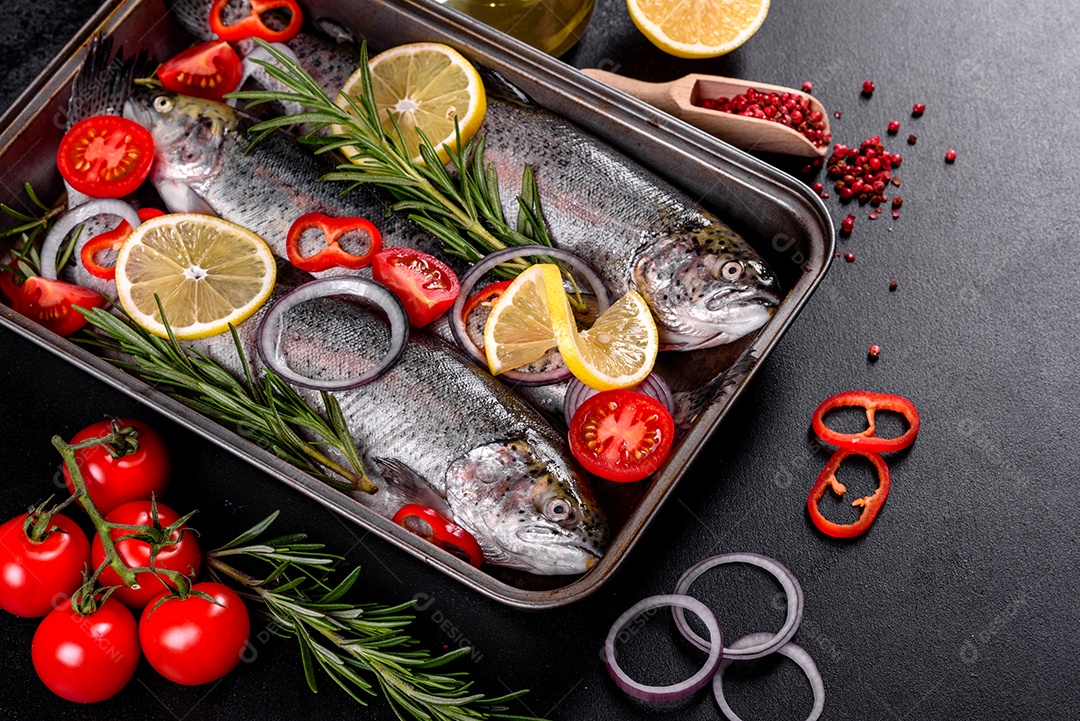 A truta crua está sobre uma tábua de pedra preta sobre uma mesa de concreto escuro. Perto estão: tomate, alecrim, limão, pimenta, cebola, sal
