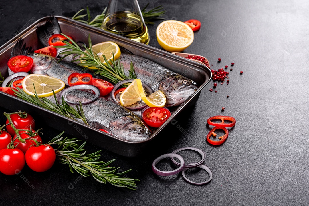 A truta crua está sobre uma tábua de pedra preta sobre uma mesa de concreto escuro. Perto estão: tomate, alecrim, limão, pimenta, cebola, sal