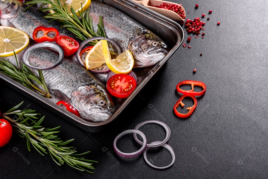 A truta crua está sobre uma tábua de pedra preta sobre uma mesa de concreto escuro. Perto estão: tomate, alecrim, limão, pimenta, cebola, sal