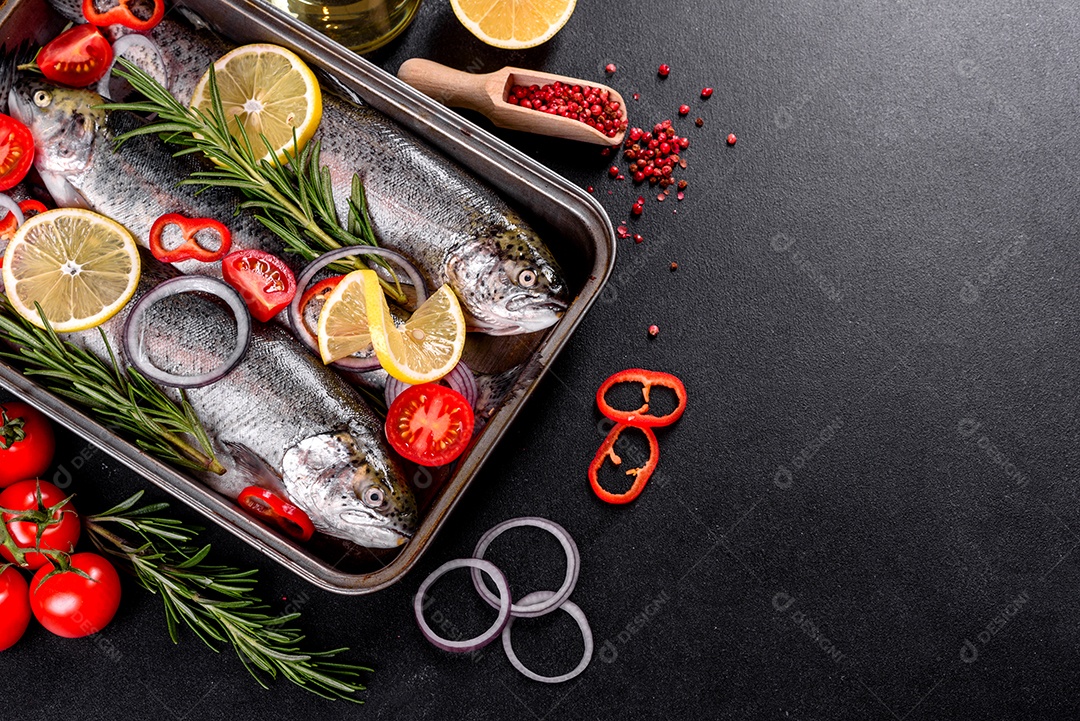 A truta crua está sobre uma tábua de pedra preta sobre uma mesa de concreto escuro. Perto estão: tomate, alecrim, limão, pimenta, cebola, sal