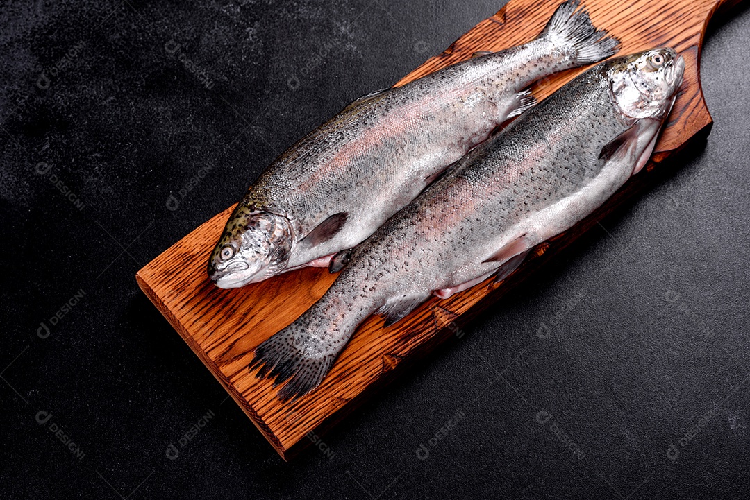 A truta crua está sobre uma tábua de pedra preta sobre uma mesa de concreto escuro. Perto estão: tomate, alecrim, limão, pimenta, cebola, sal