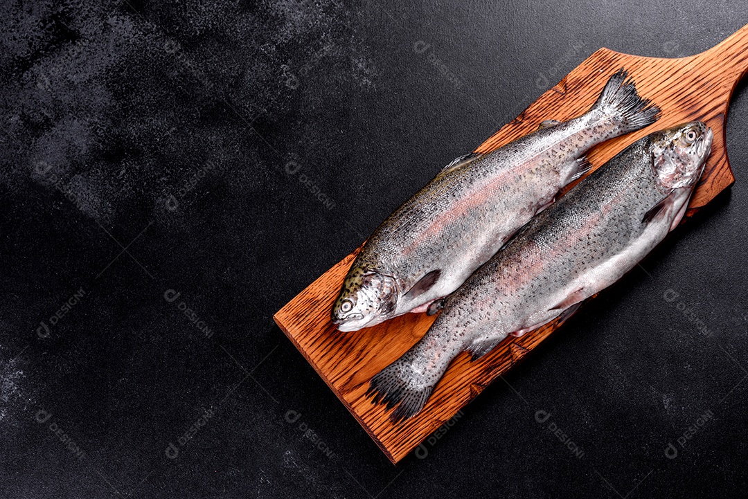 A truta crua está sobre uma tábua de pedra preta sobre uma mesa de concreto escuro. Perto estão: tomate, alecrim, limão, pimenta, cebola, sal