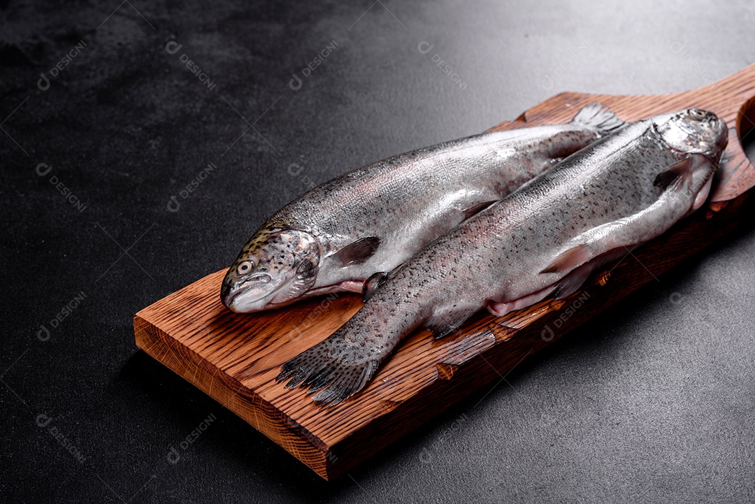 A truta crua está sobre uma tábua de pedra preta sobre uma mesa de concreto escuro. Perto estão: tomate, alecrim, limão, pimenta, cebola, sal