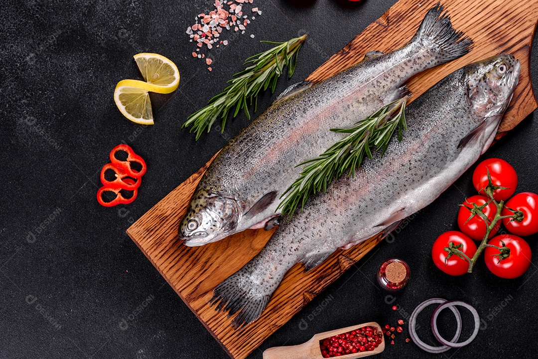 A truta crua está sobre uma tábua de pedra preta sobre uma mesa de concreto escuro. Perto estão: tomate, alecrim, limão, pimenta, cebola, sal