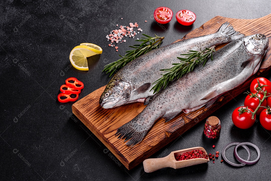 A truta crua está sobre uma tábua de pedra preta sobre uma mesa de concreto escuro. Perto estão: tomate, alecrim, limão, pimenta, cebola, sal