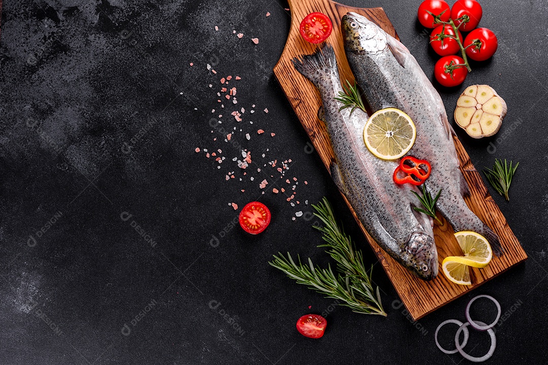 A truta crua está sobre uma tábua de pedra preta sobre uma mesa de concreto escuro. Perto estão: tomate, alecrim, limão, pimenta, cebola, sal