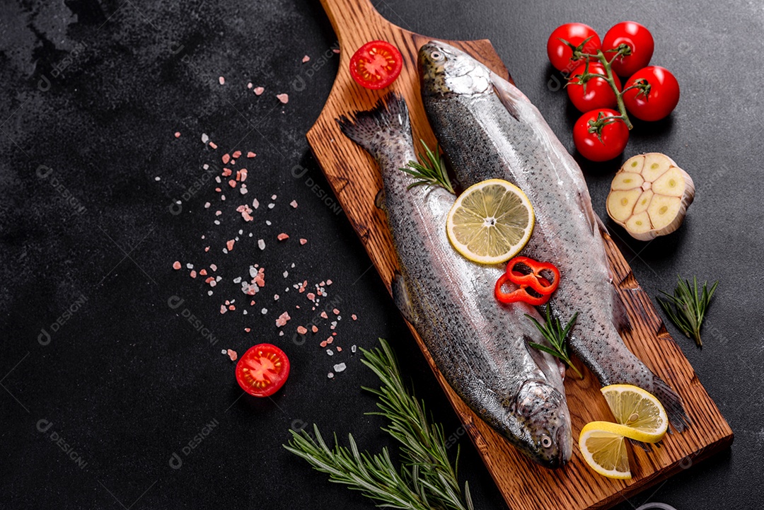 A truta crua está sobre uma tábua de pedra preta sobre uma mesa de concreto escuro. Perto estão: tomate, alecrim, limão, pimenta, cebola, sal