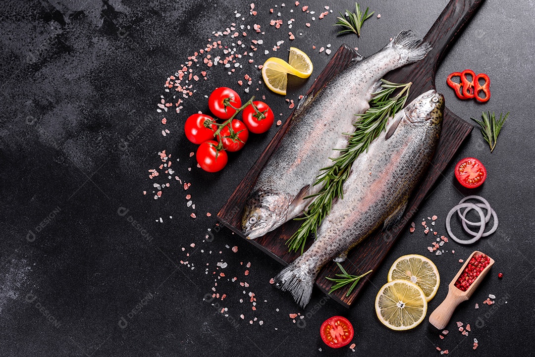 A truta crua está sobre uma tábua de pedra preta sobre uma mesa de concreto escuro. Perto estão: tomate, alecrim, limão, pimenta, cebola, sal