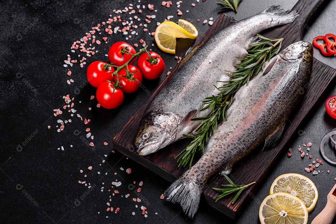 A truta crua está sobre uma tábua de pedra preta sobre uma mesa de concreto escuro. Perto estão: tomate, alecrim, limão, pimenta, cebola, sal