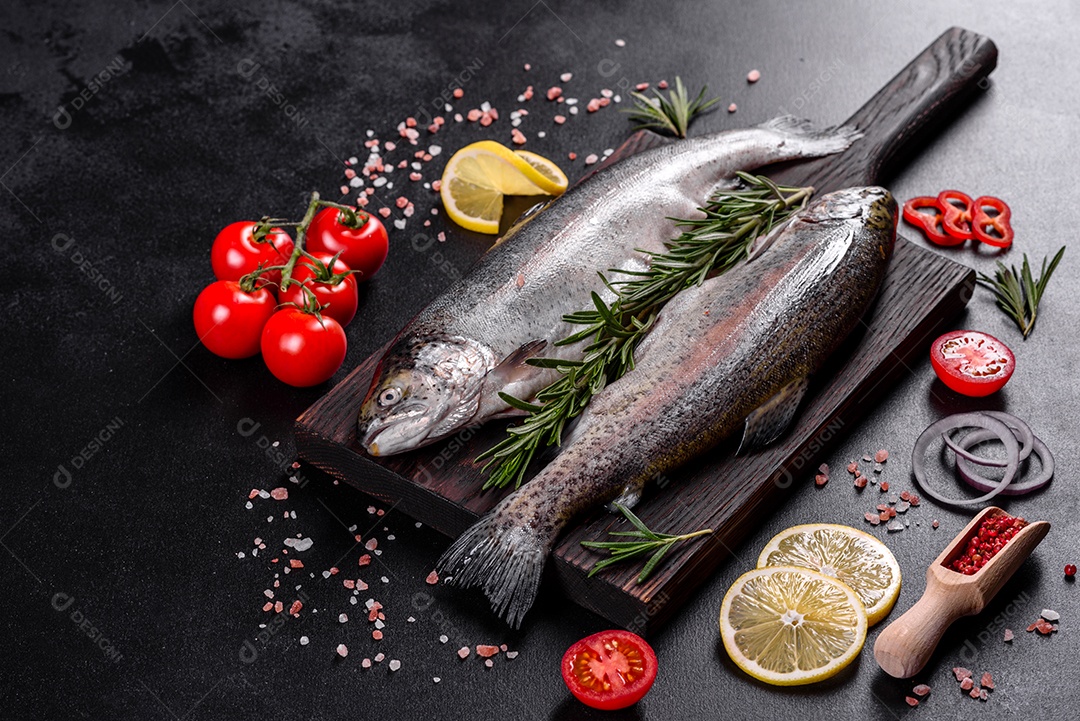 A truta crua está sobre uma tábua de pedra preta sobre uma mesa de concreto escuro. Perto estão: tomate, alecrim, limão, pimenta, cebola, sal
