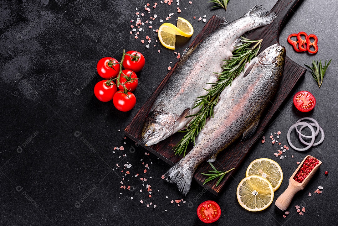 A truta crua está sobre uma tábua de pedra preta sobre uma mesa de concreto escuro. Perto estão: tomate, alecrim, limão, pimenta, cebola, sal