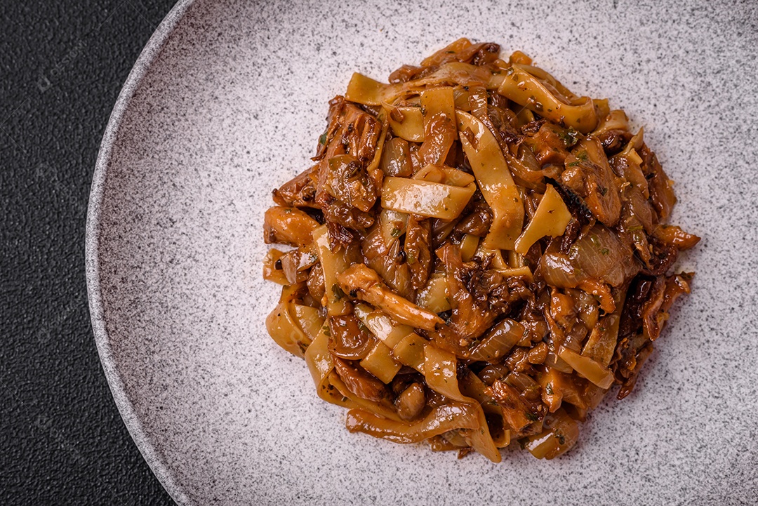 Delicioso prato de udon asiático com legumes, cogumelos, sal e especiarias numa placa cerâmica sobre um fundo escuro de concreto