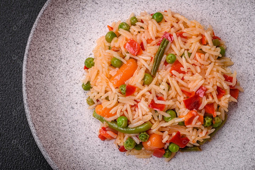Delicioso arroz cozido com legumes, pimentas, ervilhas verdes, sal, especiarias e ervas sobre um fundo escuro de concreto