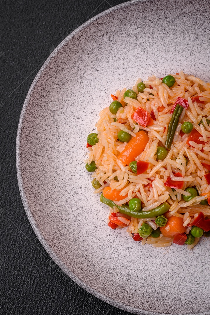 Delicioso arroz cozido com legumes, pimentas, ervilhas verdes, sal, especiarias e ervas sobre um fundo escuro de concreto