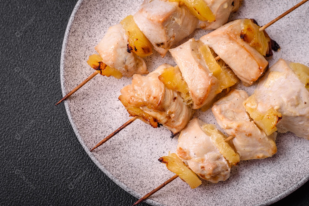 Delicioso kebab de frango ou peru com pedaços de abacaxi em espetos com sal, especiarias e ervas em um fundo escuro de concreto