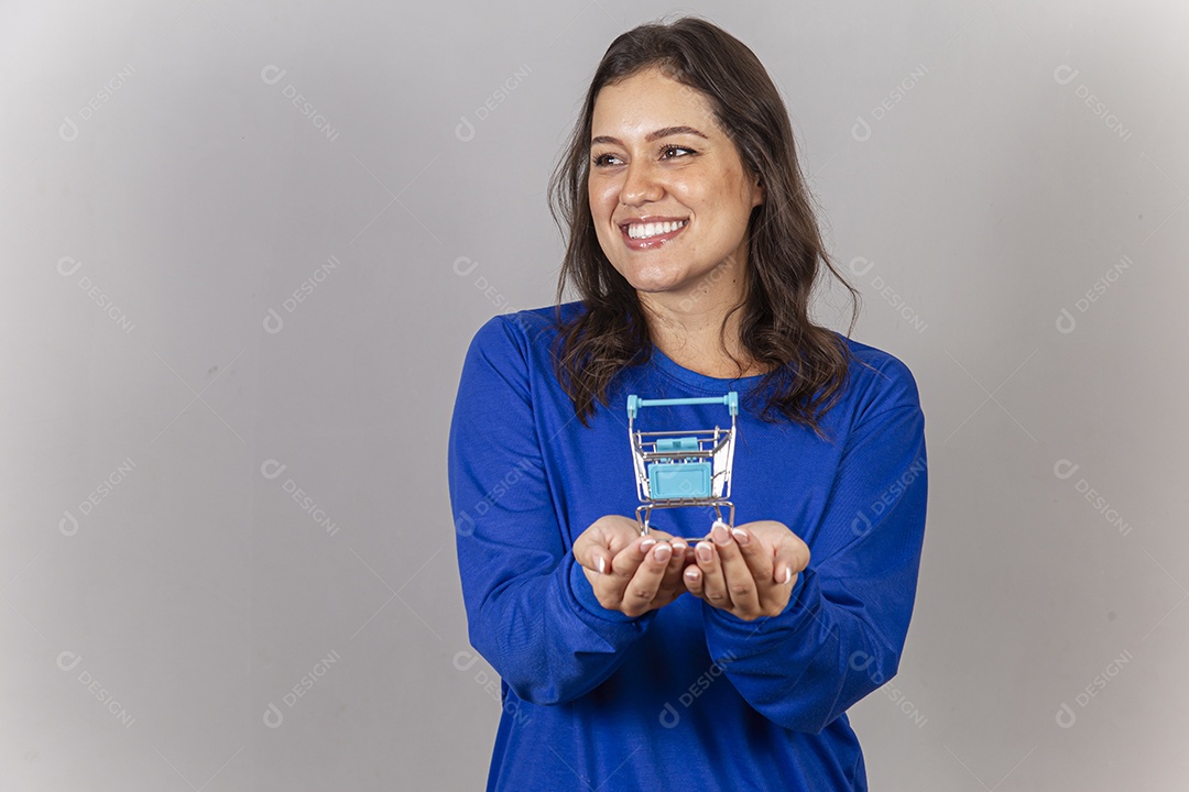 Jovem mulher sorridente com mini carrinho de supermercado nas mãos.