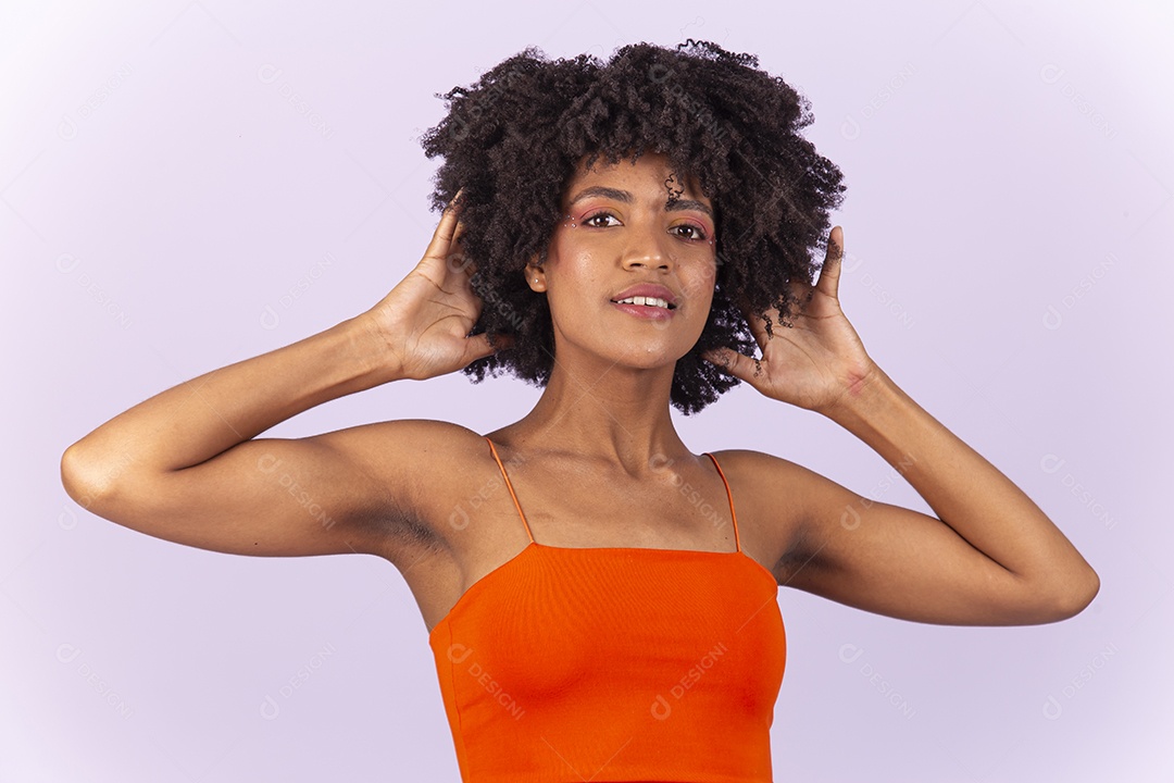 Linda jovem negra com as mãos no cabelo usando blusa laranja sobre fundo branco.