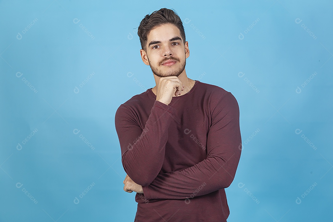 Homem jovem com mão no queixo sobre fundo isolado azul.