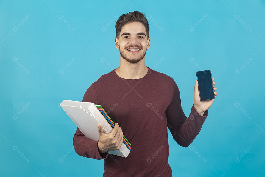 Homem jovem segurando celular e livros sobre fundo isolado azul.