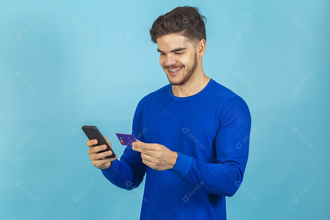 Homem segurando cartão e celular sobre um fundo isolado azul.
