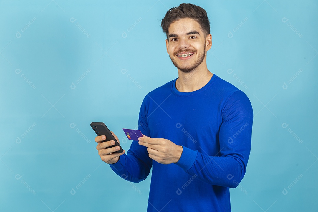 Homem segurando cartão e celular sobre um fundo isolado azul.