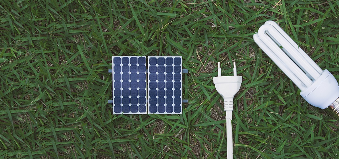 Segurando um fio de tomada e lâmpada ao lado de um painel solar fotovoltaico em fundo de grama