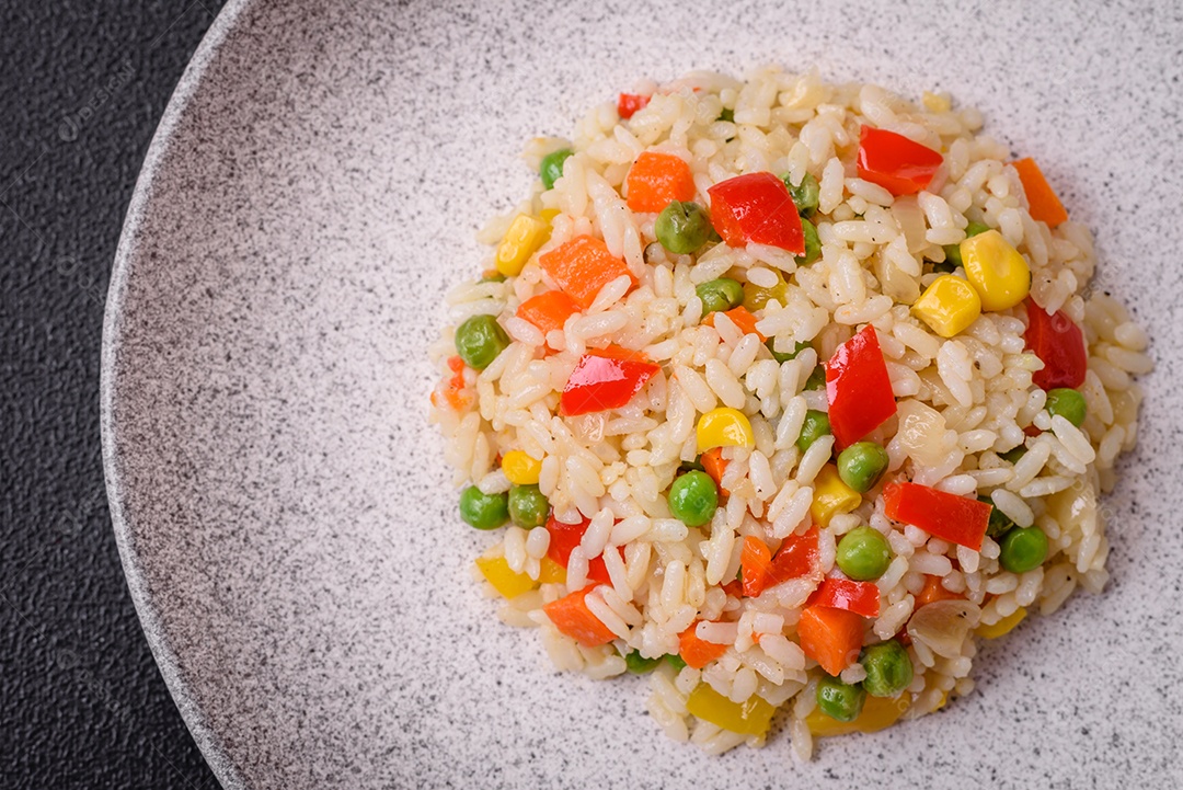 Delicioso arroz cozido branco com legumes, pimentões, cenouras, ervilhas, sal e especiarias sobre um fundo escuro de concreto