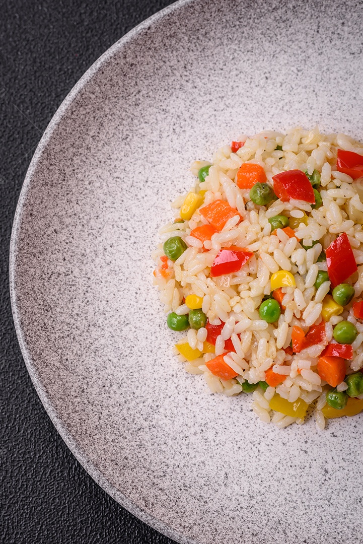 Delicioso arroz cozido branco com legumes, pimentões, cenouras, ervilhas, sal e especiarias sobre um fundo escuro de concreto