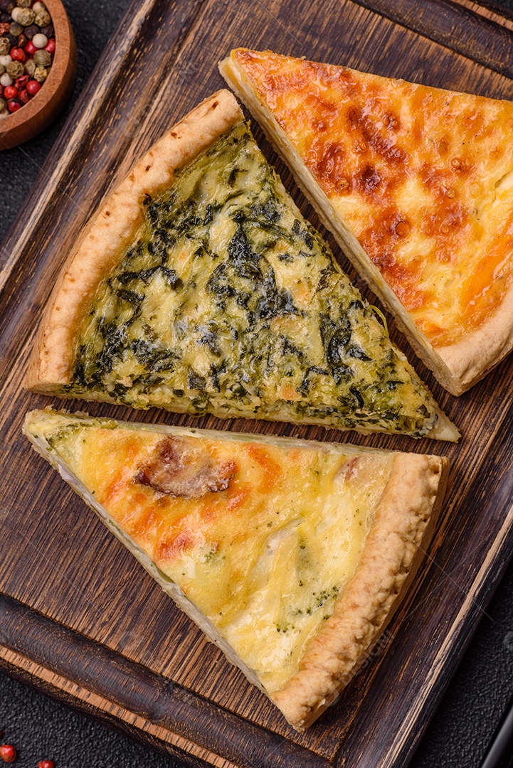 Deliciosa quiche crocante cortada em fatias com queijo, brócolis, tomate, sal, especiarias e ervas sobre um fundo escuro de concreto