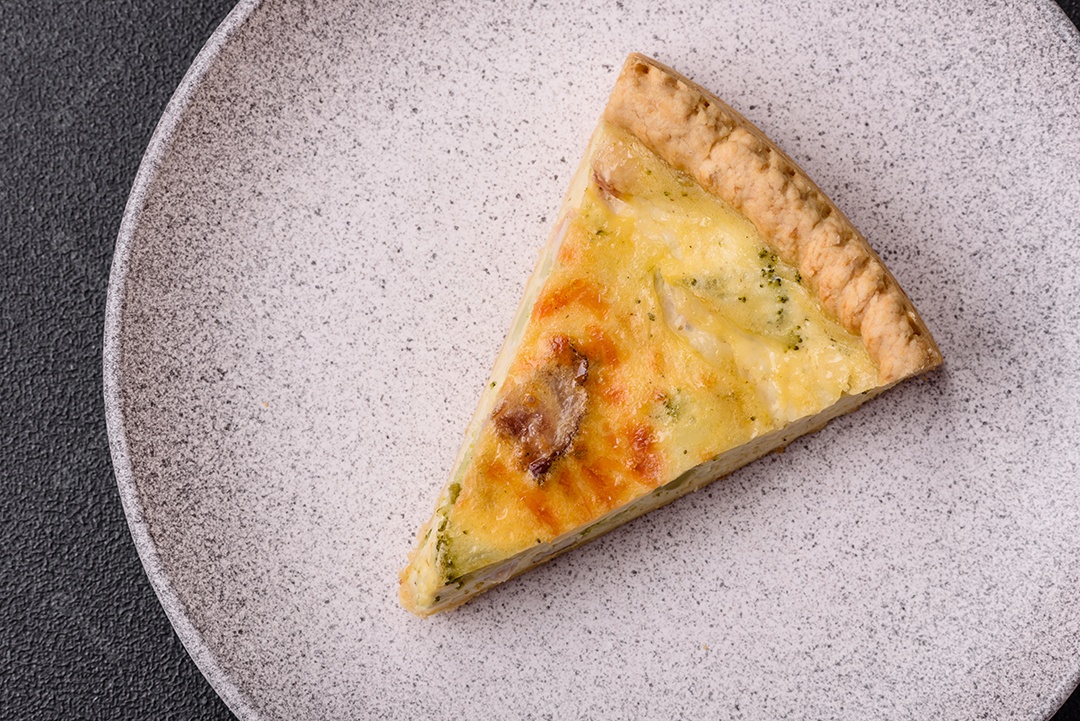 Deliciosa quiche crocante cortada em fatias com queijo, brócolis, tomate, sal, especiarias e ervas sobre um fundo escuro de concreto