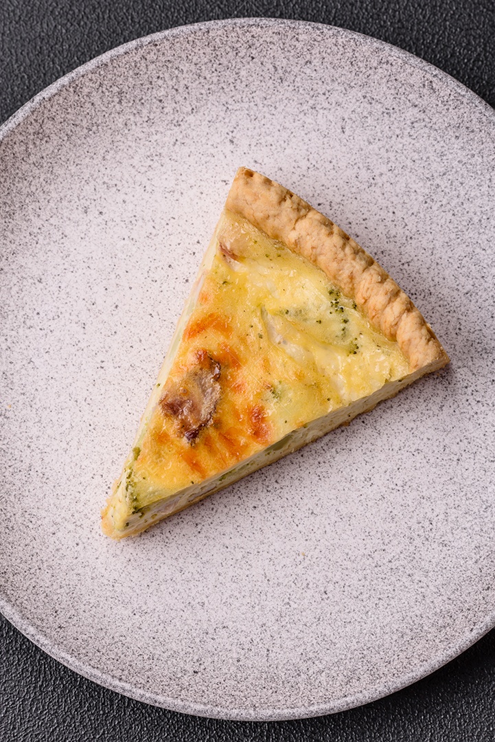 Deliciosa quiche crocante cortada em fatias com queijo, brócolis, tomate, sal, especiarias e ervas sobre um fundo escuro de concreto