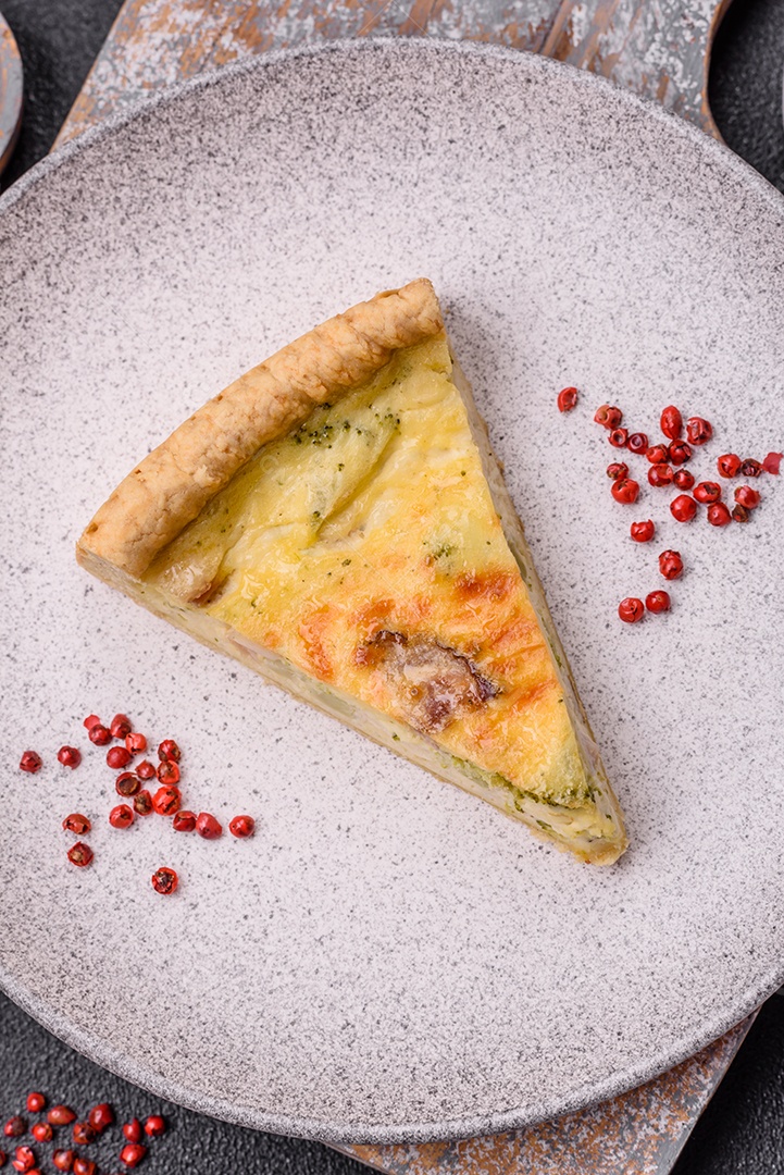 Deliciosa quiche crocante cortada em fatias com queijo, brócolis, tomate, sal, especiarias e ervas sobre um fundo escuro de concreto