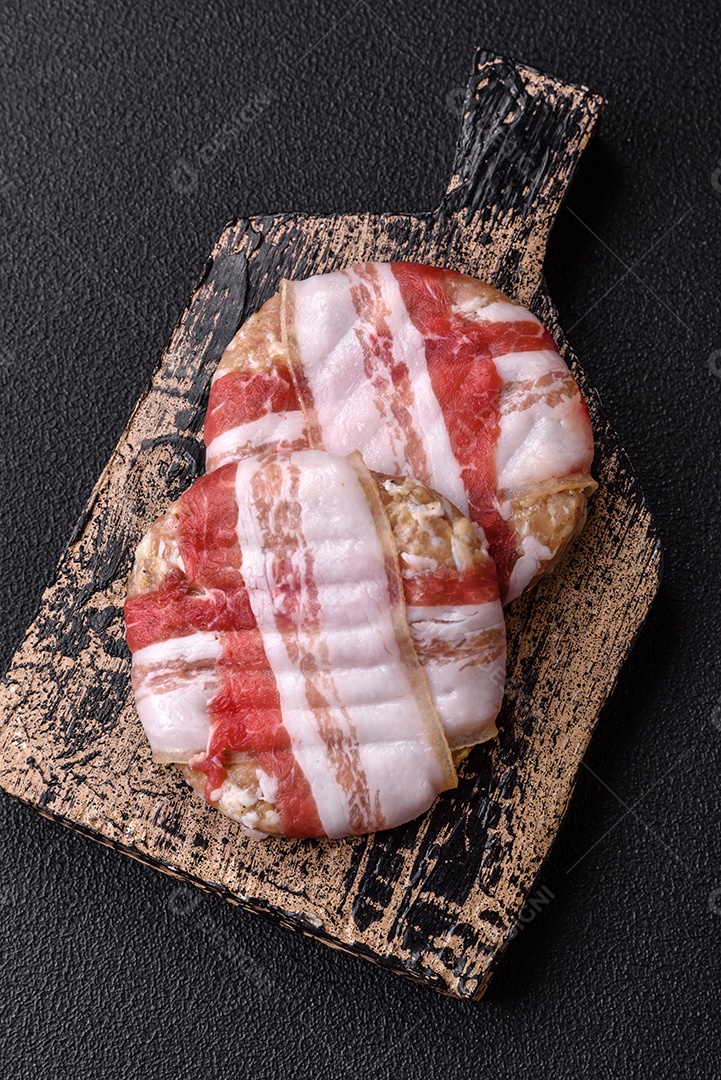 Frango picado redondo ou costeleta de porco envolta em bacon com sal, especiarias e ervas sobre um fundo escuro de concreto