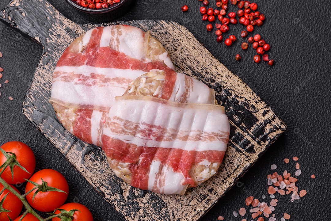 Frango picado redondo ou costeleta de porco envolta em bacon com sal, especiarias e ervas sobre um fundo escuro de concreto