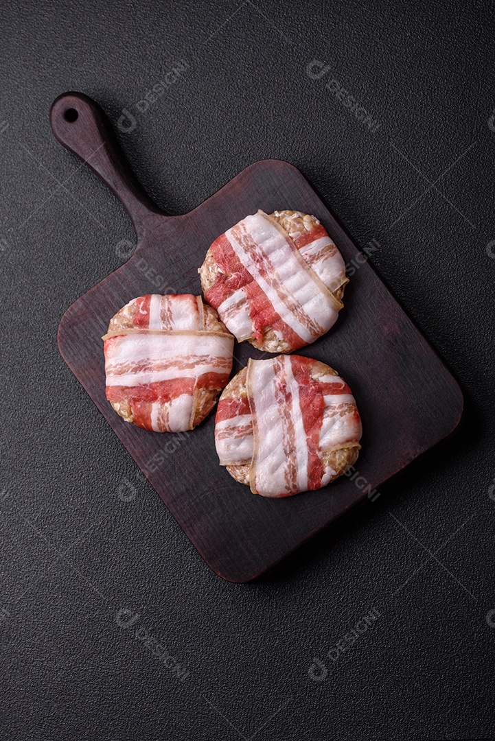Frango picado redondo ou costeleta de porco envolta em bacon com sal, especiarias e ervas sobre um fundo escuro de concreto