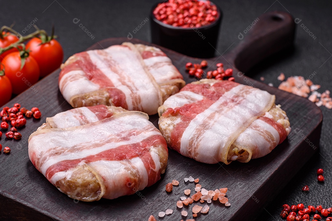Frango picado redondo ou costeleta de porco envolta em bacon com sal, especiarias e ervas sobre um fundo escuro de concreto