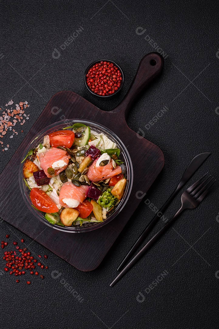 Deliciosa salada suculenta com salmão, tomate, pepino, ervas, sementes de abóbora, sal e especiarias em uma placa cerâmica sobre um fundo escuro de concreto