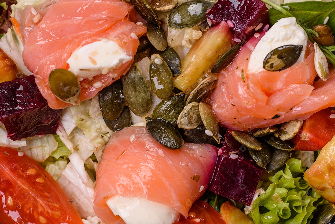 Deliciosa salada suculenta com salmão, tomate, pepino, ervas, sementes de abóbora, sal e especiarias em uma placa cerâmica sobre um fundo escuro de concreto