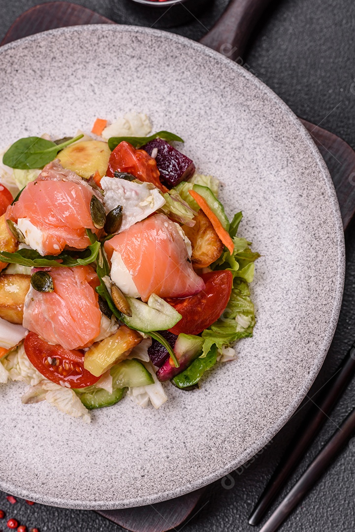 Deliciosa salada suculenta com salmão, tomate, pepino, ervas, sementes de abóbora, sal e especiarias em uma placa cerâmica sobre um fundo escuro de concreto