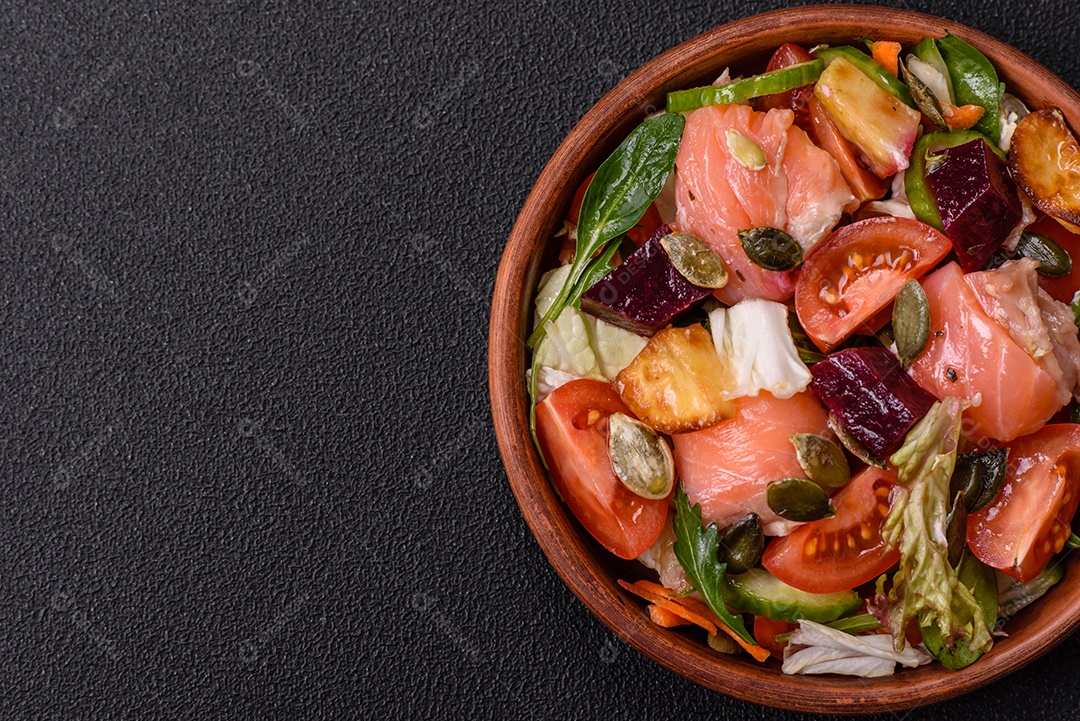 Deliciosa salada suculenta com salmão, tomate, pepino, ervas, sementes de abóbora, sal e especiarias em uma placa cerâmica sobre um fundo escuro de concreto