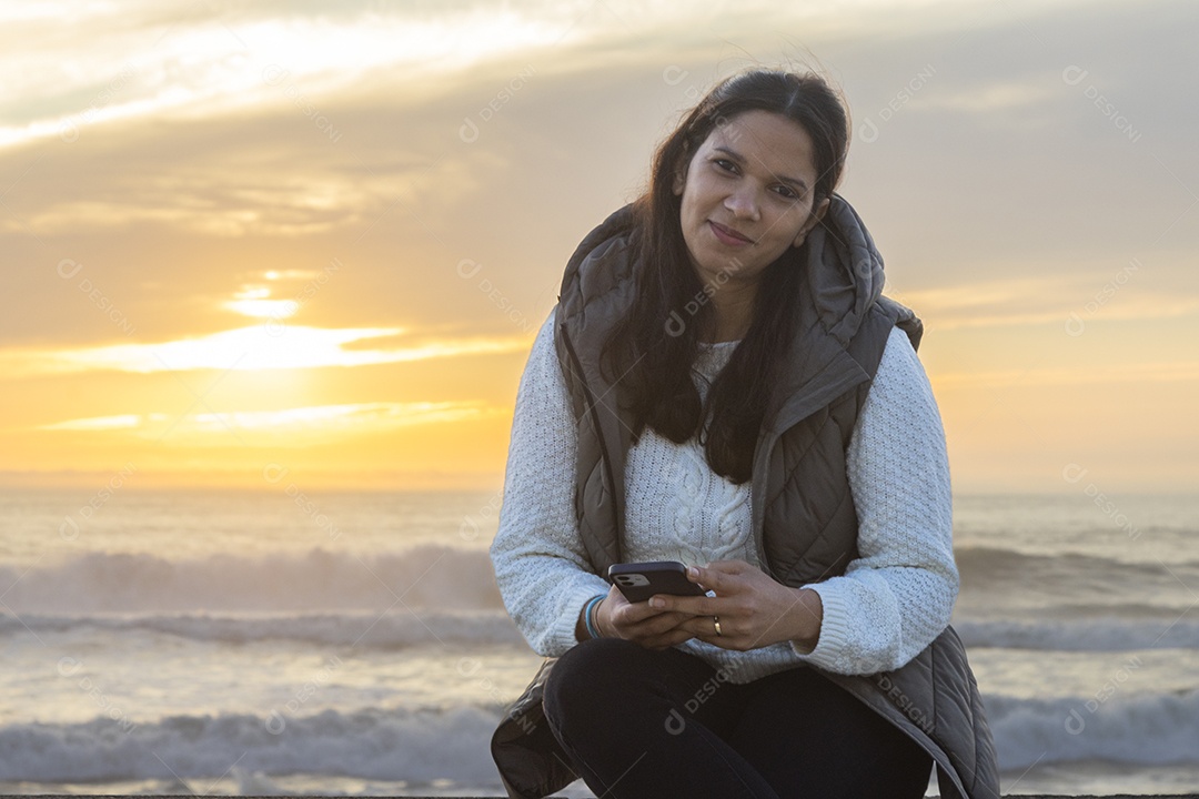 Mulher sentada segurando o celular ao pôr do sol na praia de Espinho