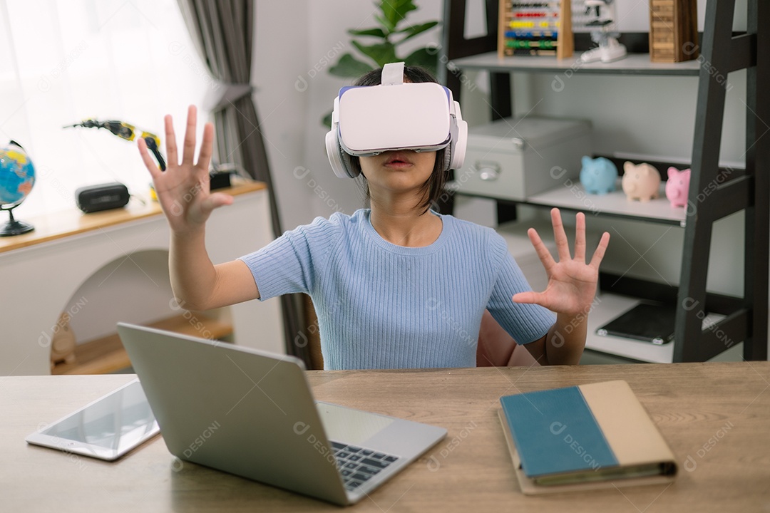 Menina usando aparelho de realidade virtual sentada em uma mesa com laptop e um livro