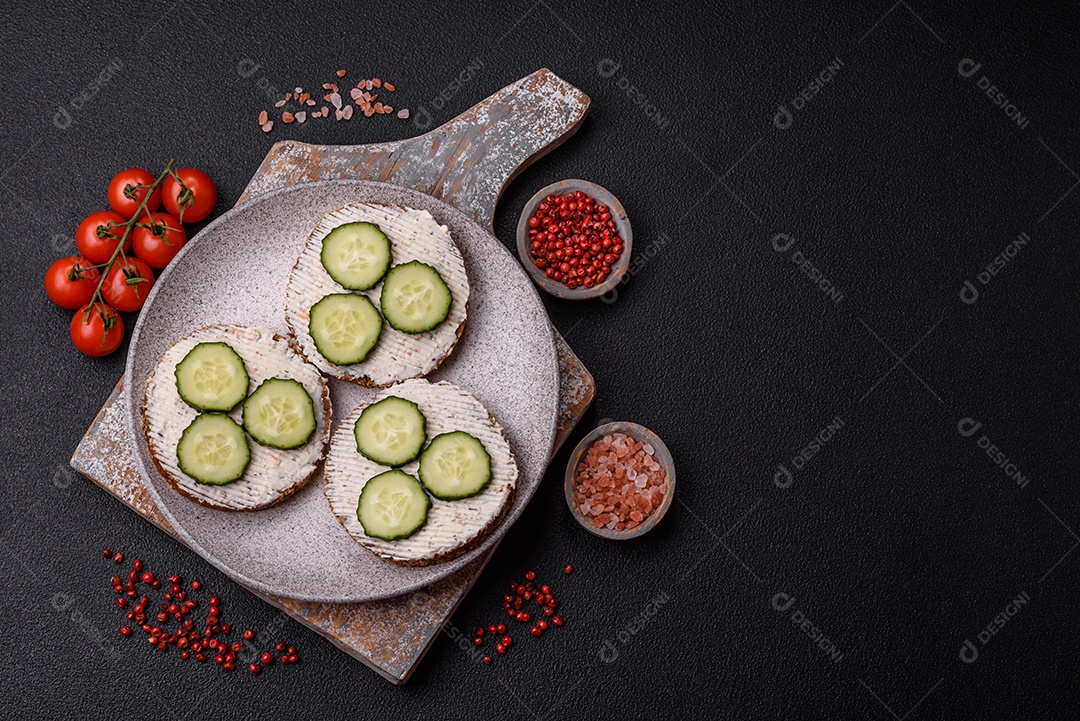 Delicioso sanduíche vegetariano com torradas grelhadas, cream cheese, pepinos e sementes e grãos sobre um fundo escuro de concreto