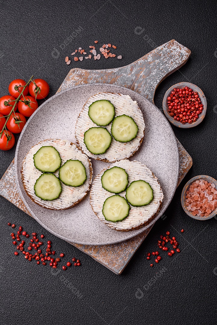 Delicioso sanduíche vegetariano com torradas grelhadas, cream cheese, pepinos e sementes e grãos sobre um fundo escuro de concreto