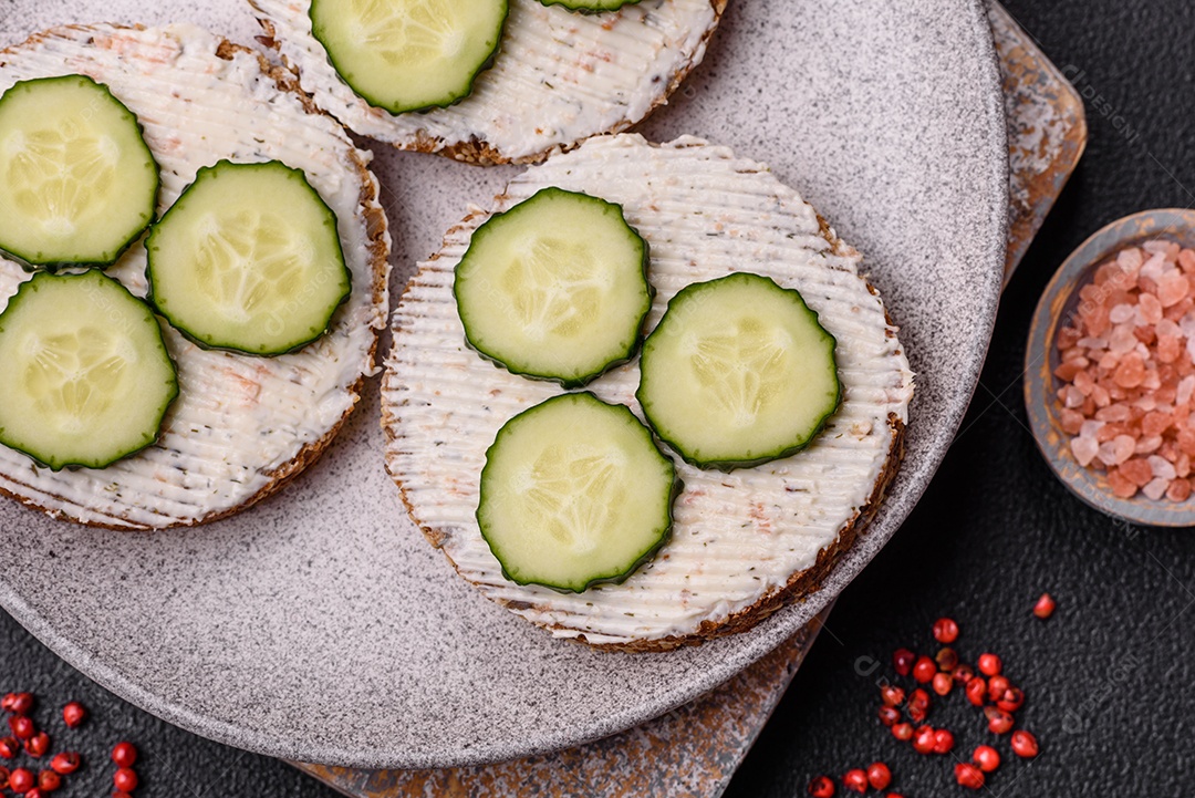 Delicioso sanduíche vegetariano com torradas grelhadas, cream cheese, pepinos e sementes e grãos sobre um fundo escuro de concreto