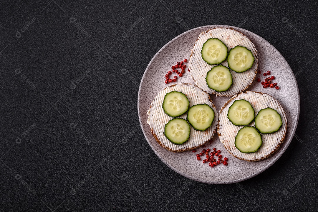 Delicioso sanduíche vegetariano com torradas grelhadas, cream cheese, pepinos e sementes e grãos sobre um fundo escuro de concreto