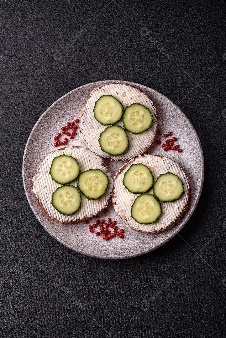 Delicioso sanduíche vegetariano com torradas grelhadas, cream cheese, pepinos e sementes e grãos sobre um fundo escuro de concreto