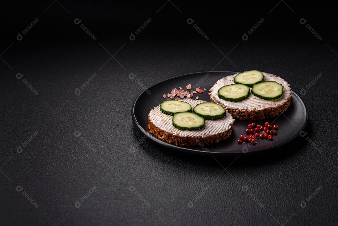 Delicioso sanduíche vegetariano com torradas grelhadas, cream cheese, pepinos e sementes e grãos sobre um fundo escuro de concreto