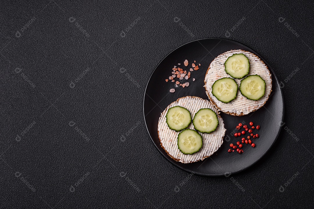 Delicioso sanduíche vegetariano com torradas grelhadas, cream cheese, pepinos e sementes e grãos sobre um fundo escuro de concreto