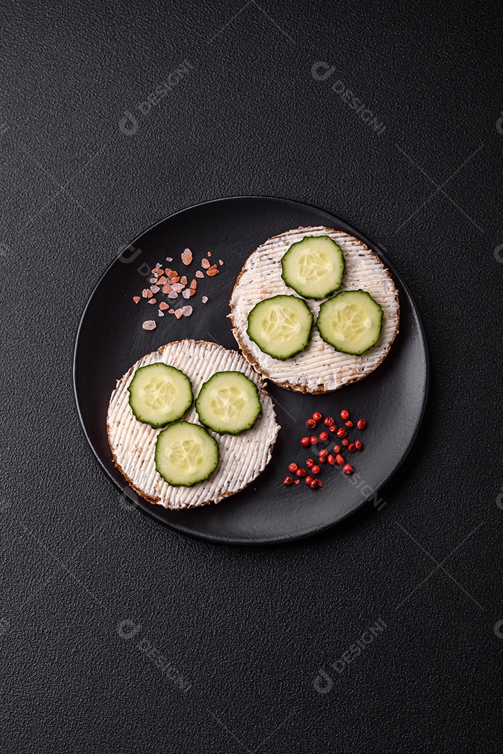 Delicioso sanduíche vegetariano com torradas grelhadas, cream cheese, pepinos e sementes e grãos sobre um fundo escuro de concreto